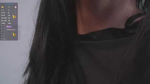 Media: Video of a woman's neck and shoulder, showcasing long, straight black hair. She wears a sheer black top, revealing her skin tone. Background includes a blurred, white wall with a digital interface displaying yellow and purple icons.