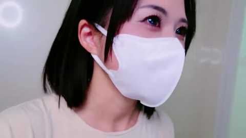 Media: Video of an Asian woman with straight, shoulder-length black hair, wearing a white face mask and a plain white top. She has light skin and is looking to the right. Background is blurry, indoor setting.