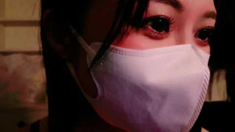 Media: A close-up video of an Asian woman wearing a white medical mask, her dark hair partially visible, with large, dark, expressive eyes. The background is blurry, suggesting an indoor setting.