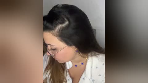 Media: Video of a young woman with long, straight black hair, wearing glasses, a white top, and a blue necklace, leaning forward, blurred background.