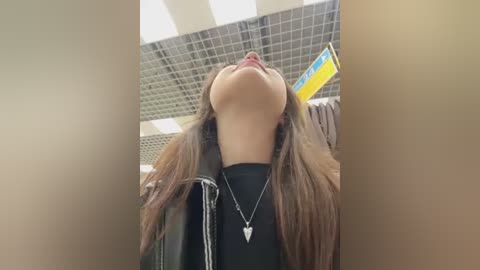 Media: Video of a young woman with long brown hair, wearing a black jacket and necklace, looking up with a serene expression in a tiled, brightly lit room.