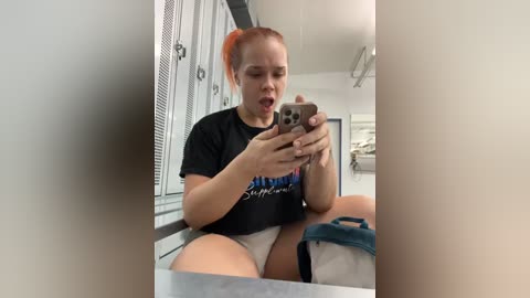 Media: Video of a young, fair-skinned woman with orange hair in a ponytail, wearing a black t-shirt and white shorts, sitting on a counter, taking a selfie in a locker room.