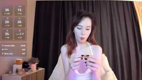 Media: Video of an Asian woman with long, dark hair, wearing a white tank top, holding a smartphone, in a dimly-lit room with black curtains and a digital clock displaying 30\u00b0F.