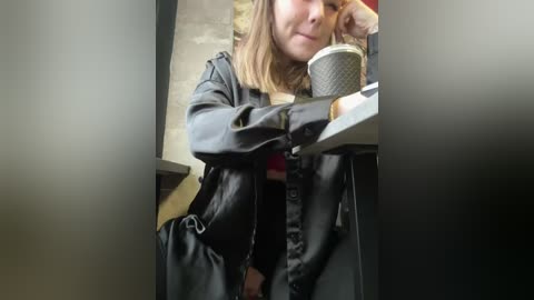 Media: Video of a woman with shoulder-length brown hair, wearing a black leather jacket, holding a black coffee cup, sitting in a dimly lit cafe with stone walls.