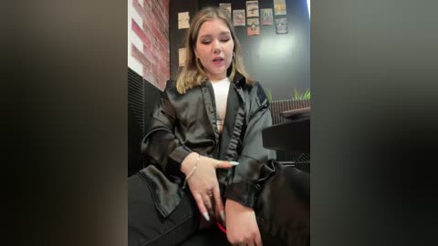 Media: Video of a young woman with shoulder-length blonde hair, wearing a black leather jacket and white top, sitting on a black chair, adjusting a black dog's leash. Background includes a brick wall, framed photos, and a dark room.