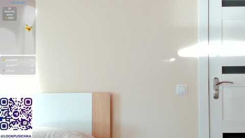 Media: A video of a minimalist bedroom with a white wall, a light wood headboard, a QR code in the lower left corner, and a white door with a silver handle on the right.