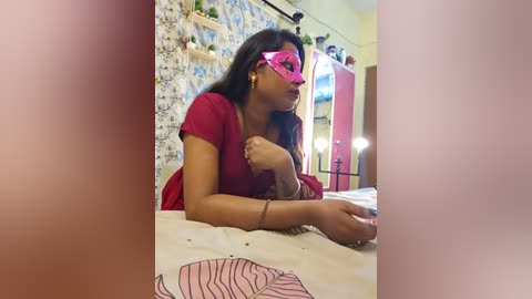 Media: Video of a South Asian woman with medium brown skin, long black hair, wearing a pink mask, red dress, and bracelets, lying on a bed with floral bedding, surrounded by a colorful, patterned wall.