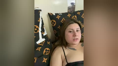 Media: A video of a plus-sized woman with light skin and brown hair, wearing a black tank top, lying on a bed with black sheets adorned with orange Louis Vuitton patterns.