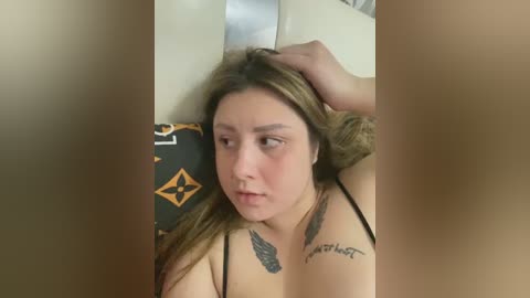 Media: Video of a young woman with long brown hair, fair skin, and multiple tattoos, including feathers, resting her head on a pillow with a black and yellow pattern. She looks pensive.