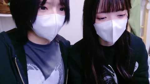 Media: Video of two East Asian women wearing white surgical masks, one in a gray T-shirt, the other in a black jacket, sitting indoors.