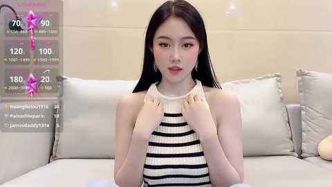 Media: A video of an Asian woman with long black hair and fair skin, wearing a striped sleeveless top, sitting on a bed with light pillows and beige walls.