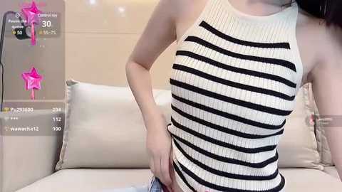 Media: Video of a slim Asian woman in a sleeveless, ribbed, black-and-white striped tank top, standing in a modern, beige-themed room with a white couch and cushions.