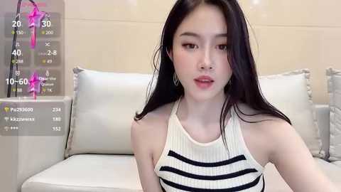 Media: Video of a young Asian woman with long black hair, wearing a sleeveless white top with black stripes, sitting on a white couch in a modern living room.