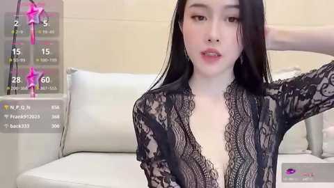 Media: Video of an East Asian woman with long black hair, wearing a black lace dress, sitting on a white sofa, looking into the camera.