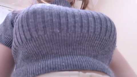 Media: A video of a woman wearing a tight, ribbed gray turtleneck sweater that accentuates her large breasts. Her fair skin and blonde hair are visible. The background is out of focus, suggesting an indoor setting.