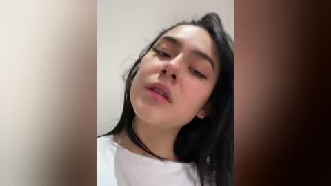 Media: Video of a young woman with light skin and long black hair, wearing a white top, leaning against a beige wall, eyes closed, looking relaxed.