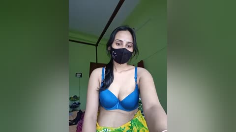 Media: Video of a South Asian woman with medium build and long black hair, wearing a blue bra, black face mask, and yellow floral skirt in a dimly lit, green-walled room.