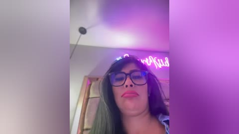 Media: Video of a woman with long black hair, wearing black-rimmed glasses and red lipstick, captured in a dimly lit room with purple neon lighting and a door in the background.