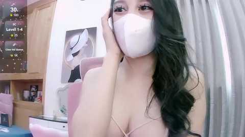 Media: Video of a young Asian woman with long black hair, wearing a white face mask, revealing ample cleavage in a revealing top. Background includes a pink chair and a white wall with a framed art piece.