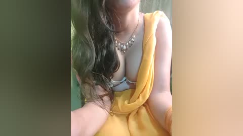 Media: A video of a fair-skinned woman with long, wavy brown hair, wearing a yellow robe, revealing a white bra with lace trim. She's indoors, near a beige wall, with a silver necklace.