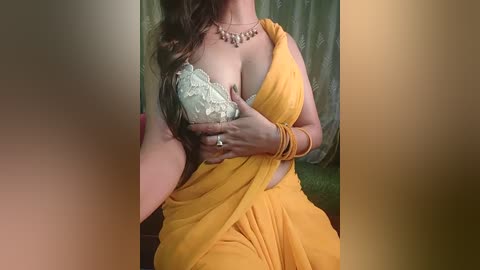 Media: Video of a woman with medium skin tone, wearing a yellow saree with a white lace bra visible. She has long, dark hair, a gold necklace, and multiple gold bangles. Background features green curtains.