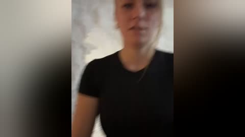 Media: A blurry video of a woman with light skin and blonde hair, wearing a black t-shirt, standing indoors against a blurred background.