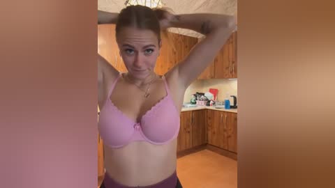 Media: Video of a light-skinned woman with fair skin and brown hair in a pink bra, standing in a wooden kitchen with cabinets, countertop, and utensils.