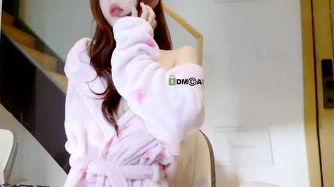 Media: Video of a young woman with long, wavy brown hair, wearing a pink, fluffy robe with a green \"EMCOAL\" logo, sitting at a desk, holding a phone to her ear.