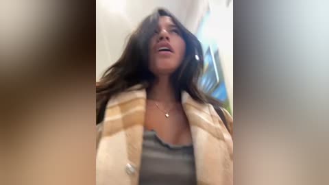 Media: A video of a woman with long brown hair, wearing a gray tank top and a beige and white plaid scarf, standing indoors with blurred background.