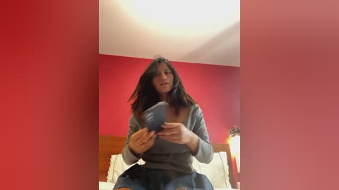 Media: A video of a woman with long dark hair, wearing a gray sweater, sitting on a bed in a room with red walls, holding a blue cup.