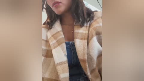 Media: Video of a woman with medium-brown skin and shoulder-length brown hair, wearing a beige and white plaid blanket over a blue denim shirt, in a blurred, indoor setting.