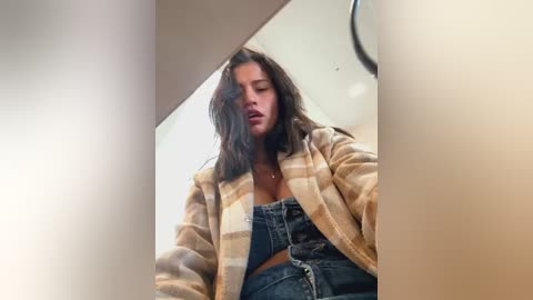 Media: Video of a young woman with wavy, dark hair, wearing a beige plaid cardigan and denim shorts, sitting in a dimly lit room with a round clock on the wall.