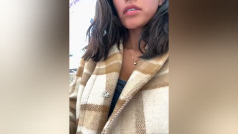 Media: A video of a young woman with medium-length, wavy, dark brown hair wearing a beige and brown plaid blanket over a black top, showcasing her lightly tanned skin. The background is blurred.