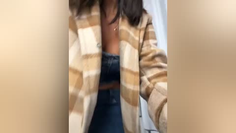 Media: Video of a woman wearing a beige and brown plaid flannel shirt over a dark blue bra, with her hands resting on her waist, partially visible in a blurred, indoor setting.