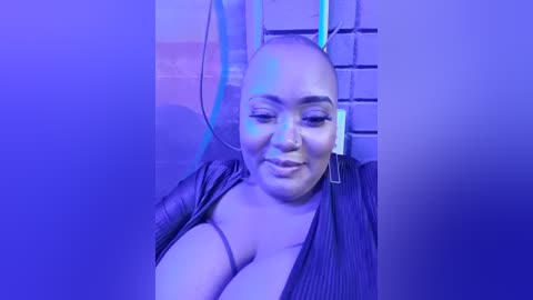 Media: A video of a smiling, bald Black woman with medium skin tone, wearing a black jacket, sitting in a brick-wall room, with blue lighting and a blurred background.