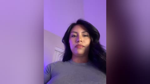 Media: Video of a young woman with long, dark hair, wearing a gray t-shirt, standing against a purple-lit wall.