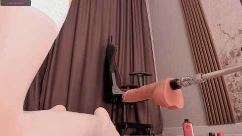 Media: Video of a realistic sex doll with a large, erect penis, seated on a gaming chair, with brown curtains and a cracked wall in the background.