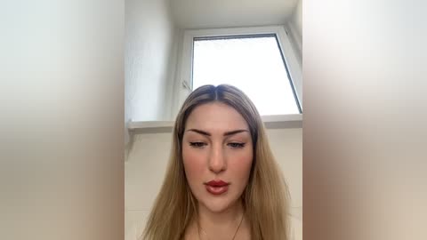 Media: A video of a young woman with long, straight blonde hair and fair skin, wearing red lipstick and a white top, standing in a bright attic room with white walls and a skylight.