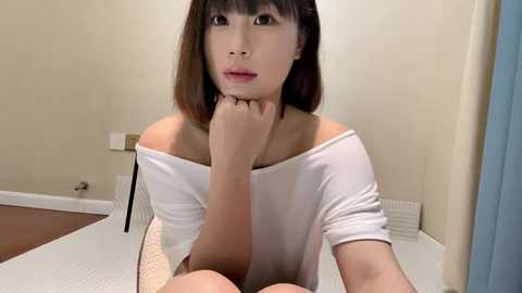 Media: A video of an Asian woman with straight, shoulder-length brown hair, wearing a white off-shoulder top, sitting on a bed with white sheets, in a plain beige room.