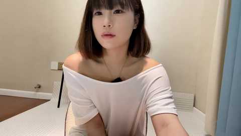 Media: Video of an East Asian woman with short brown hair, wearing a white off-shoulder top, sitting on a white bed in a beige room.