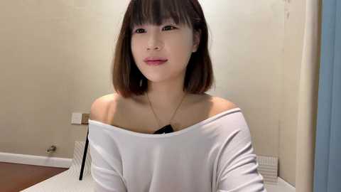 Media: Video of a young East Asian woman with straight brown hair and bangs, wearing a white off-shoulder top, standing in a beige-walled room with a radiator and door frame visible.