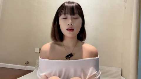 Media: Video of an Asian woman with shoulder-length black hair, wearing a white off-shoulder top, sitting in a minimalist room with beige walls and a radiator.