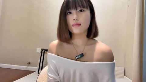 Media: Video of an East Asian woman with straight, shoulder-length dark hair, wearing a white off-the-shoulder top, standing in a beige room with a wooden floor and white baseboard.