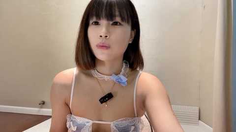 Media: Video of a young Asian woman with shoulder-length brown hair, wearing a lace bra and blue bow choker, in a beige-walled room with white radiator.