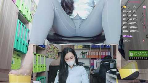 Media: A video of an Asian woman with long black hair wearing a white mask and light grey pants, sitting in an office with shelves filled with green and yellow binders.