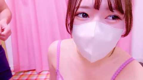 Media: Video of a young Asian woman with short brown hair, wearing a white face mask and a pink and white striped spaghetti strap top, against a pink background.