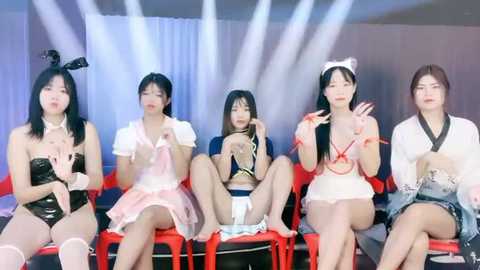 Media: Video of five Asian women in revealing, anime-inspired costumes, sitting on red chairs, under bright lights, creating a playful and risqu\u00e9 atmosphere.