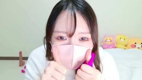 Media: A video of an Asian woman with long black hair, wearing a pink surgical mask, holding a pink vibrator, against a plain beige wall with stuffed toys.