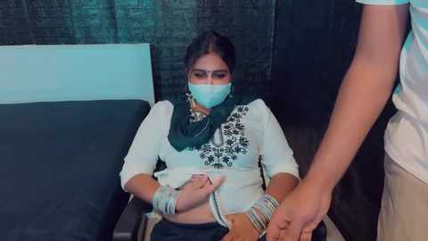 Media: Video of a woman with a mask, dark hair, wearing a white top and green scarf, being restrained by a man's hands in a medical room with dark walls.