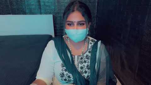 Media: Video of a South Asian woman with dark skin, wearing a traditional white and green saree with intricate embroidery, a blue mask, and a bindi. She sits on a dark bed with a white pillow against a dark wall.